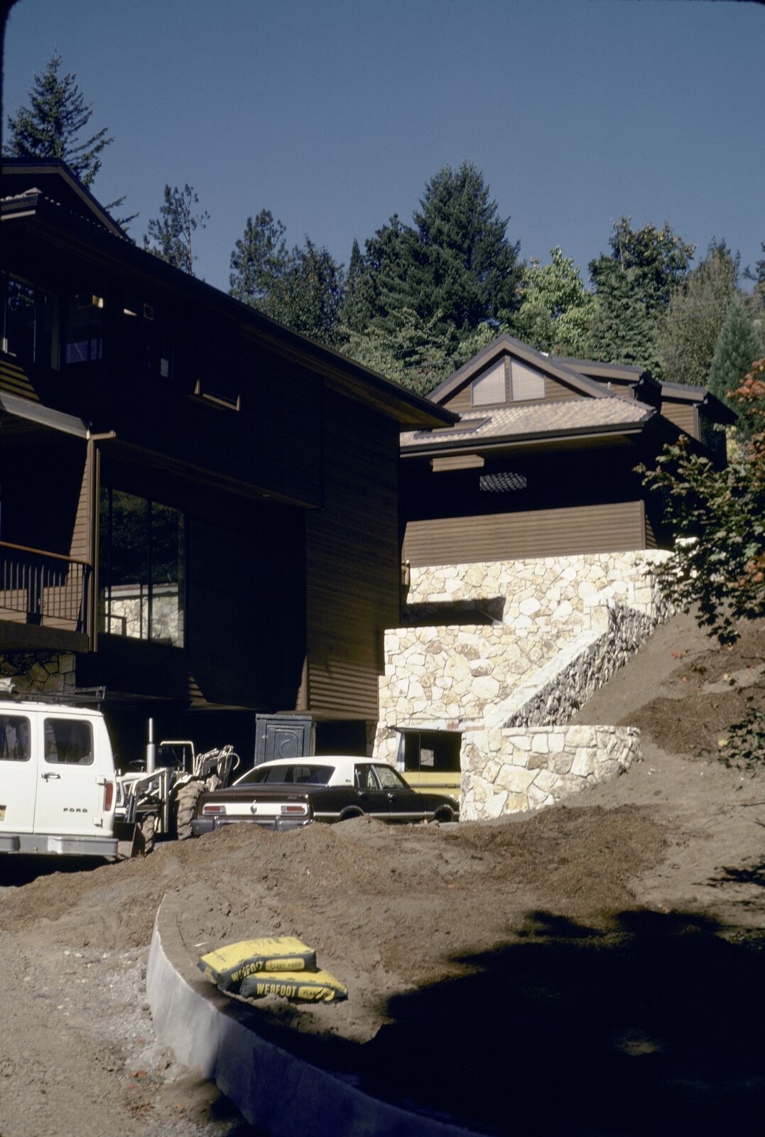 The Zimel House during construction: dark-stained siding, a cantilevered upper floor with balconies, a gabled wing on a stone base, firs behind, a van, a loader, and bags of Webfoot plant food in the drive.